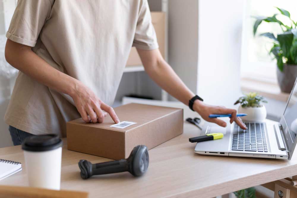 Order Processing and Package Preparation Person preparing a package for shipment while entering order information on a laptop at a packing workstation.